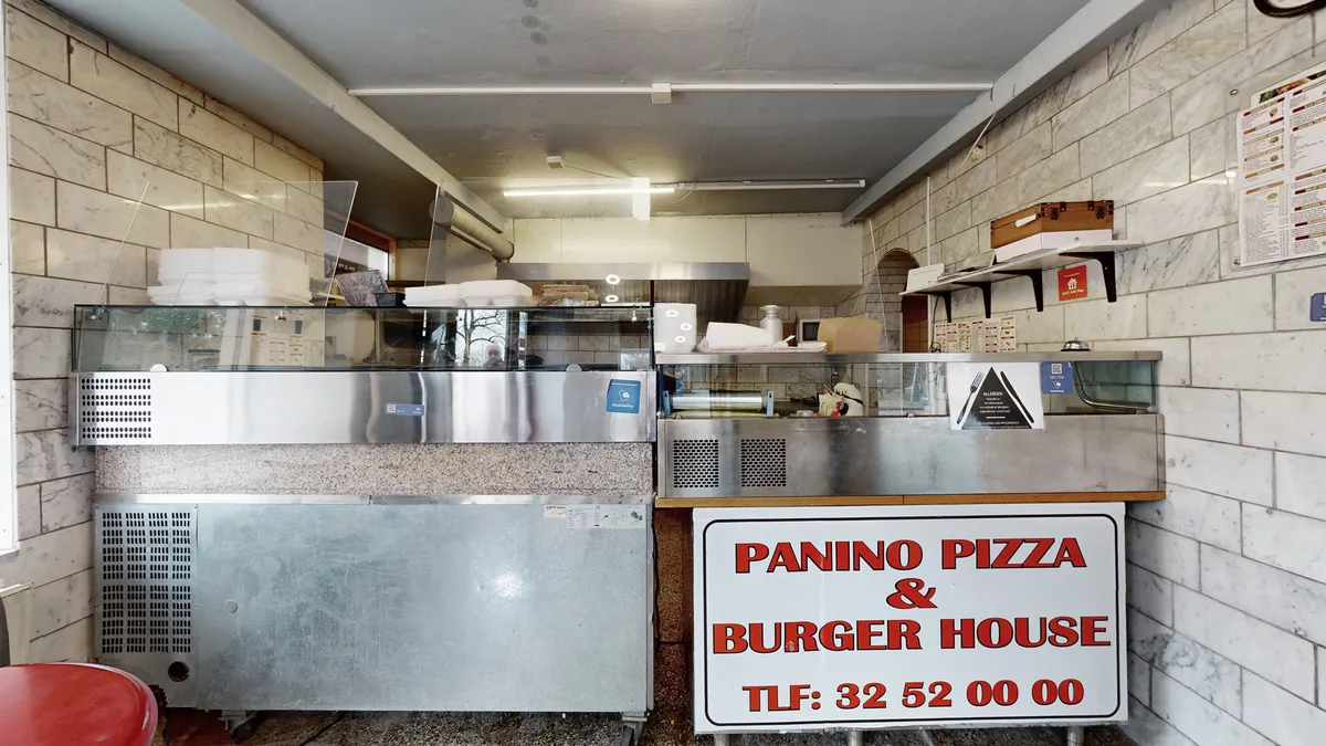 Pizzarestaurant med køkken med flere bøfsandwicher og pizzaer på skufferne, samt skilte med navnet Panino Pizza & Burger