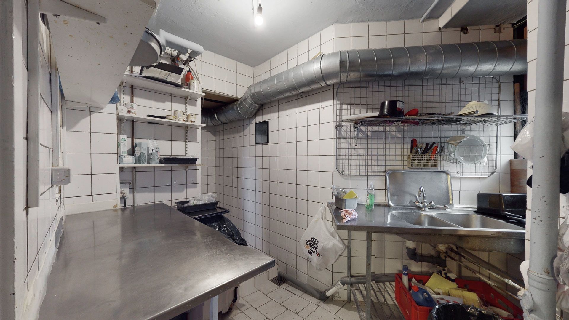 En køkkenrum med en stor metalbord og en sink samt en hængende hætte over en komfur. Der er et rør fastgjort til loftet og