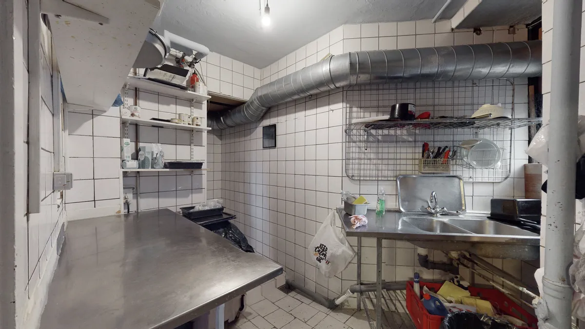 En køkkenrum med en stor metalbord og en sink samt en hængende hætte over en komfur. Der er et rør fastgjort til loftet og