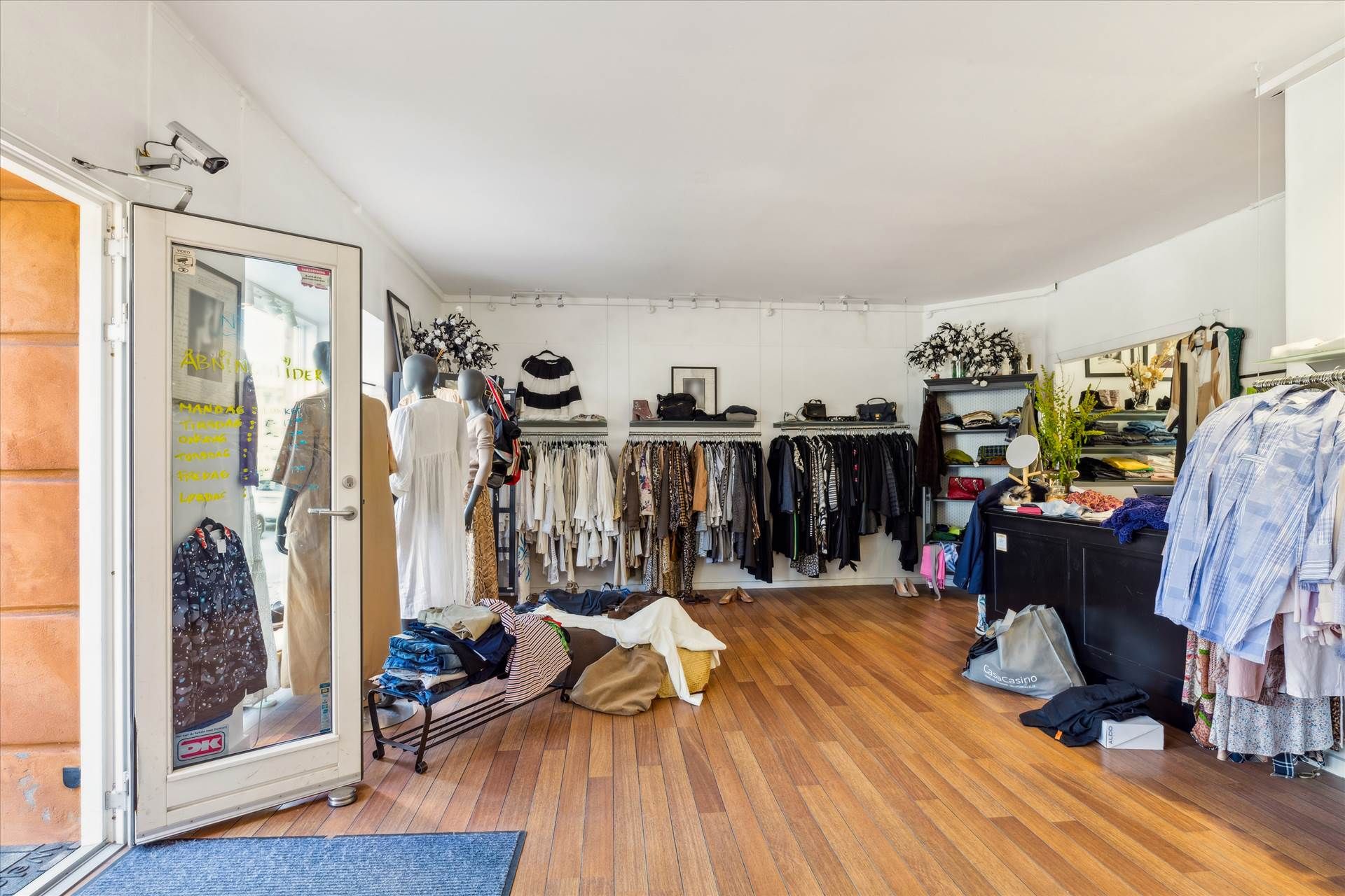 Inde i en klædeskab er der tøj på hylder og tøjvogne samt to mannequiner med tøj. Der er flere hvide vægge med plakater, og