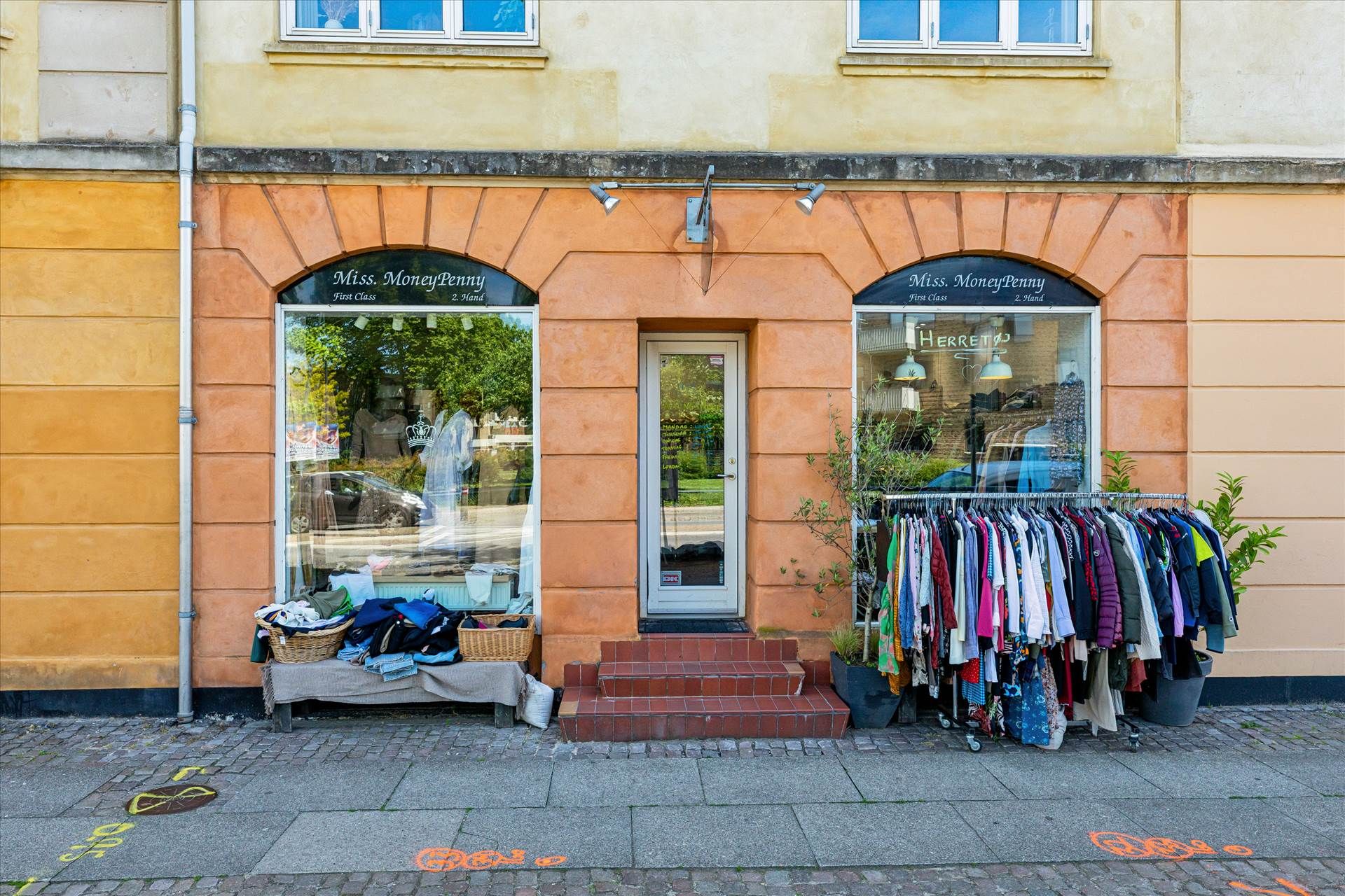 Køb og salg af klæder og anden brugt tøj i butikken Miss. Money Penny. Sorte sko og tøjvogne med tøj udenfor butikken