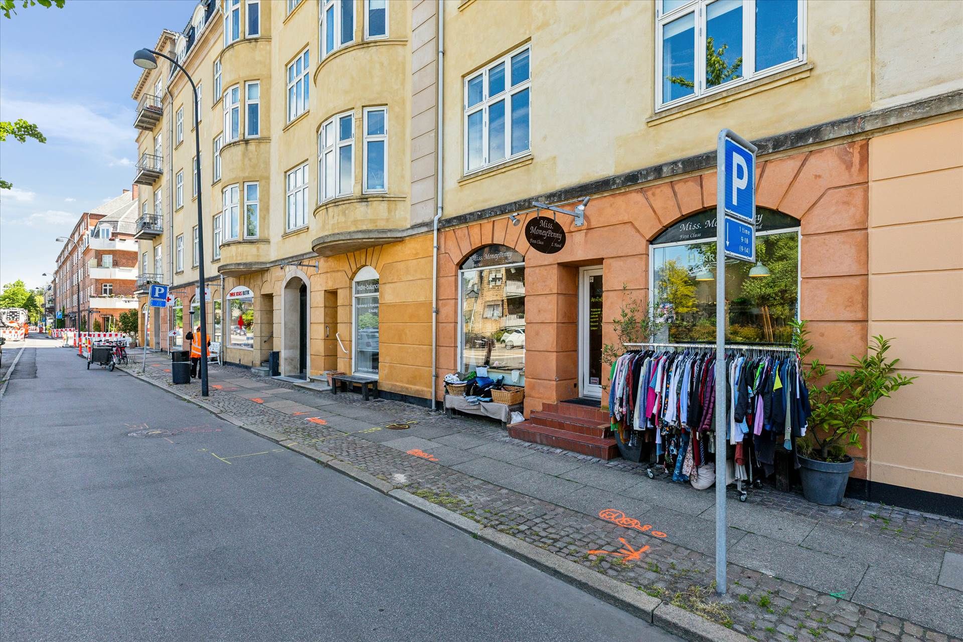 En tom gade med butikker, hvor den ene har tøj udstillet på gaden, parkeringsskilt og en søjle med skrift