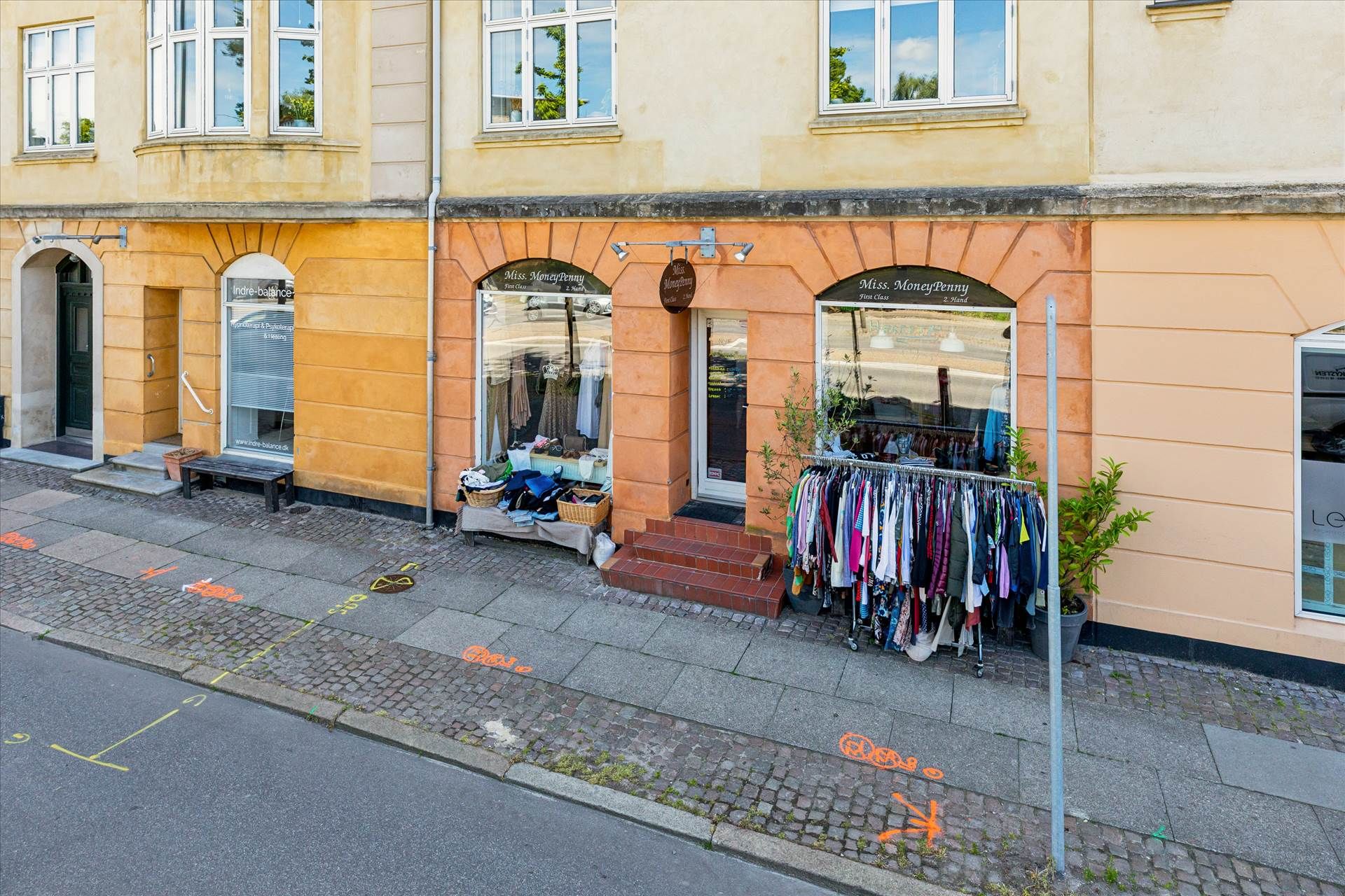 Foto af butikslokaler med tøj til salg på hylderne og en plante i potter på siden af gaden.