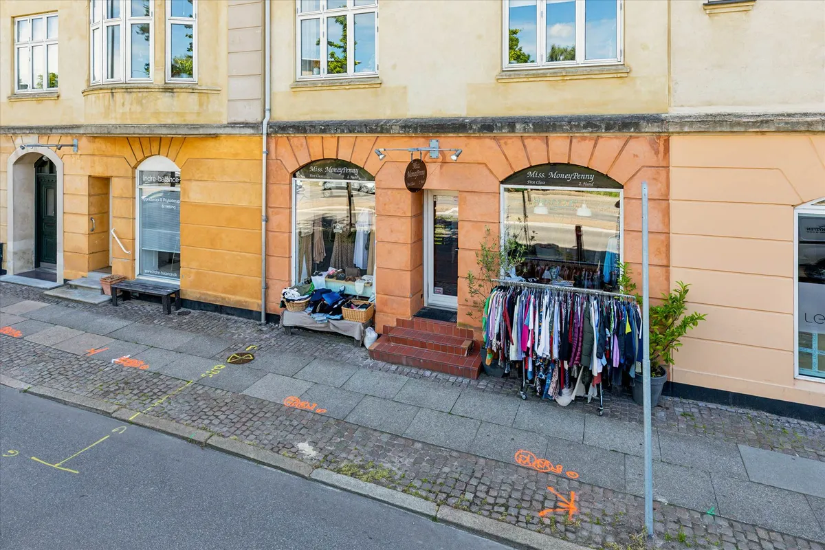 Foto af butikslokaler med tøj til salg på hylderne og en plante i potter på siden af gaden.