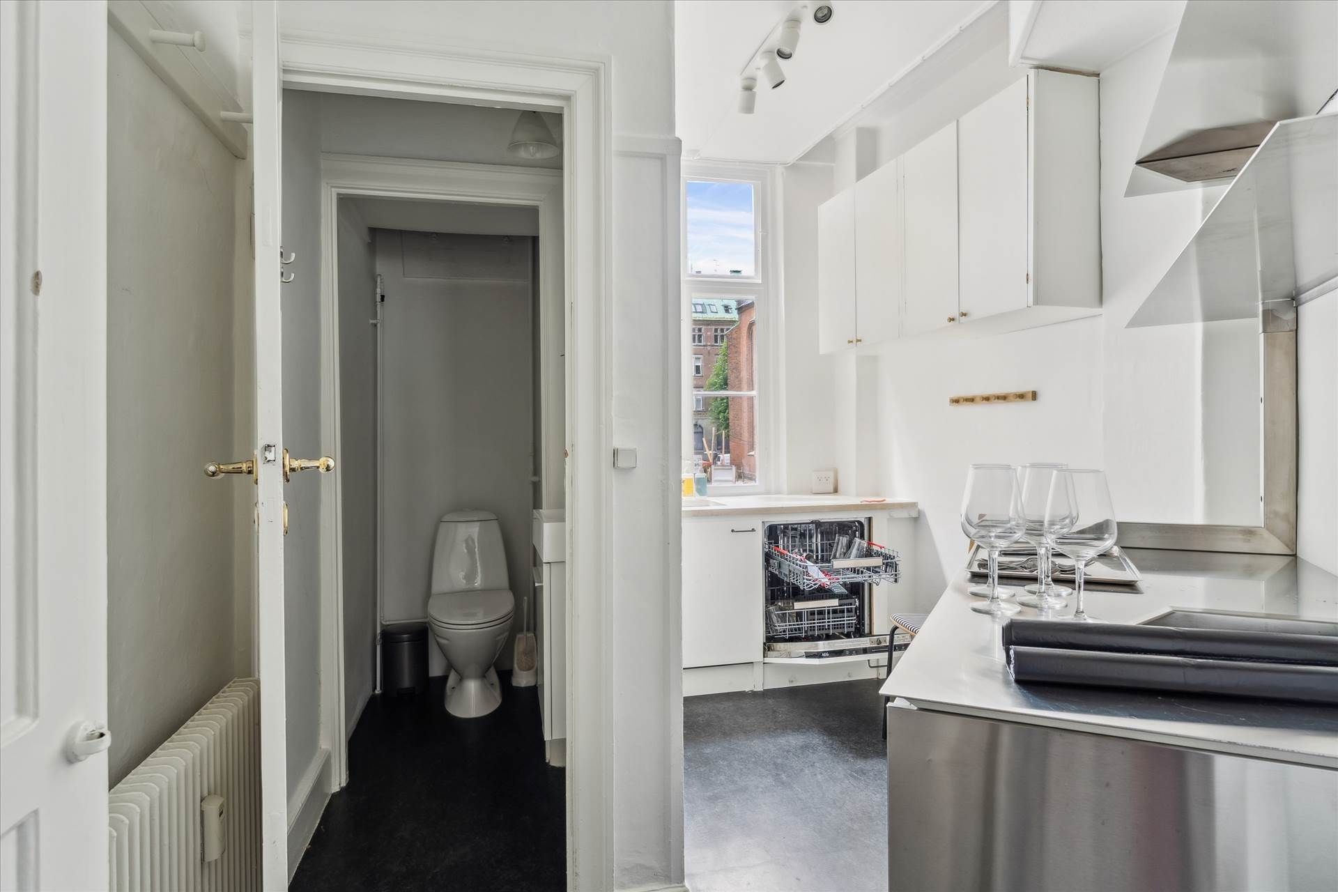 Et minimalistisk design med hvide vægge og en stor toilet i et lille rum, der leder til et køkken med rustfrit stål og en