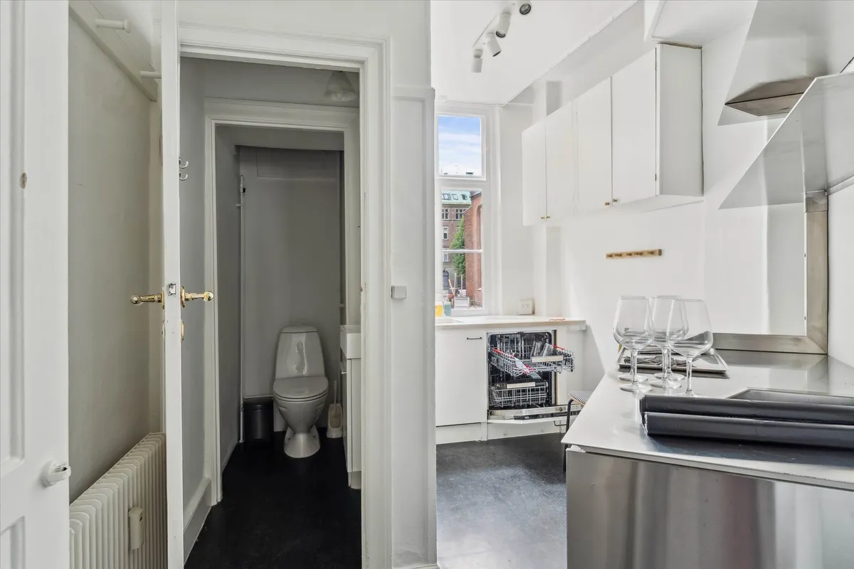 Et minimalistisk design med hvide vægge og en stor toilet i et lille rum, der leder til et køkken med rustfrit stål og en