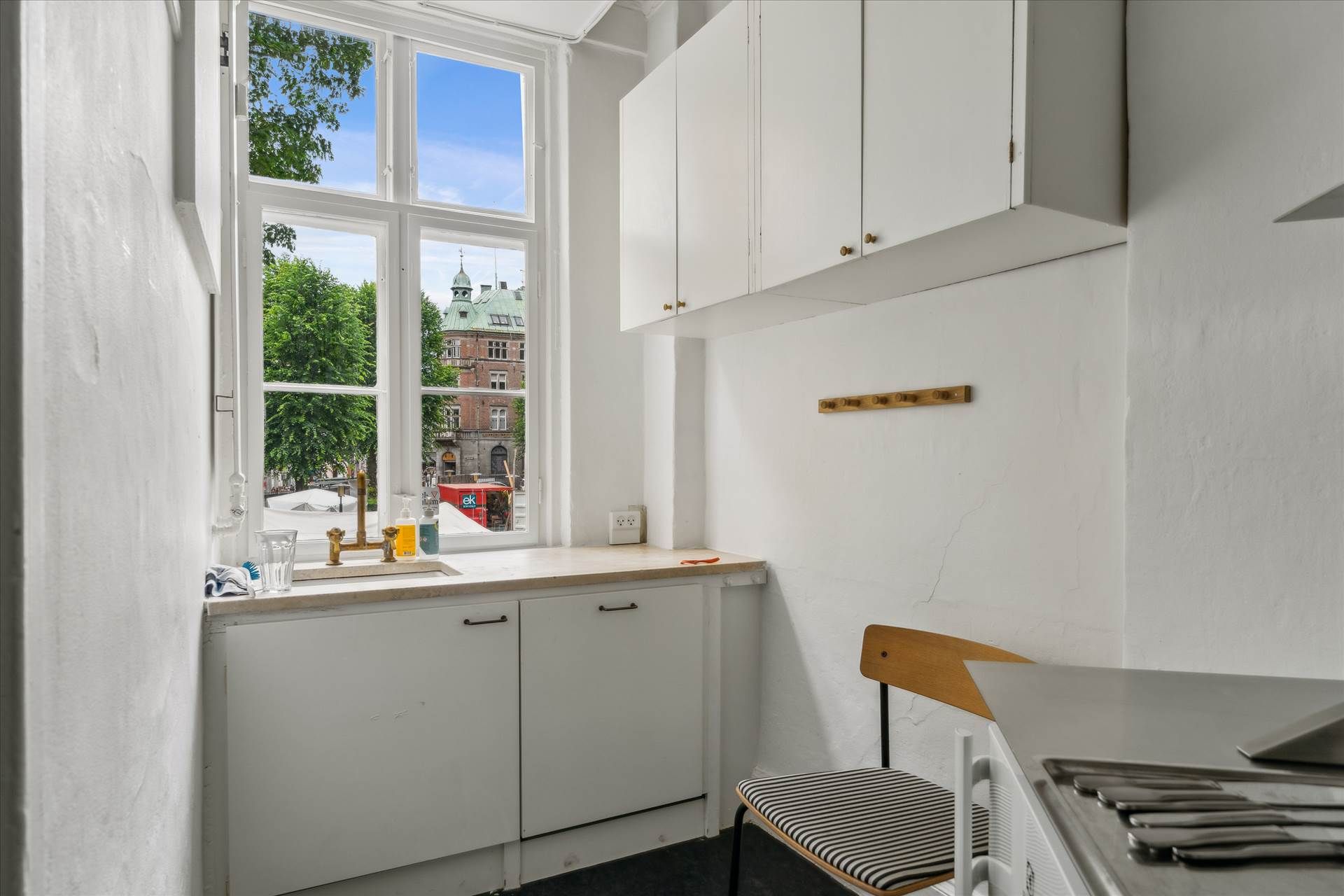 Lyst lejemål med køkken, hvidt køkkeninventar og køkkenvæg med loftskappe, og udsigt til grønne træer og bygninger i