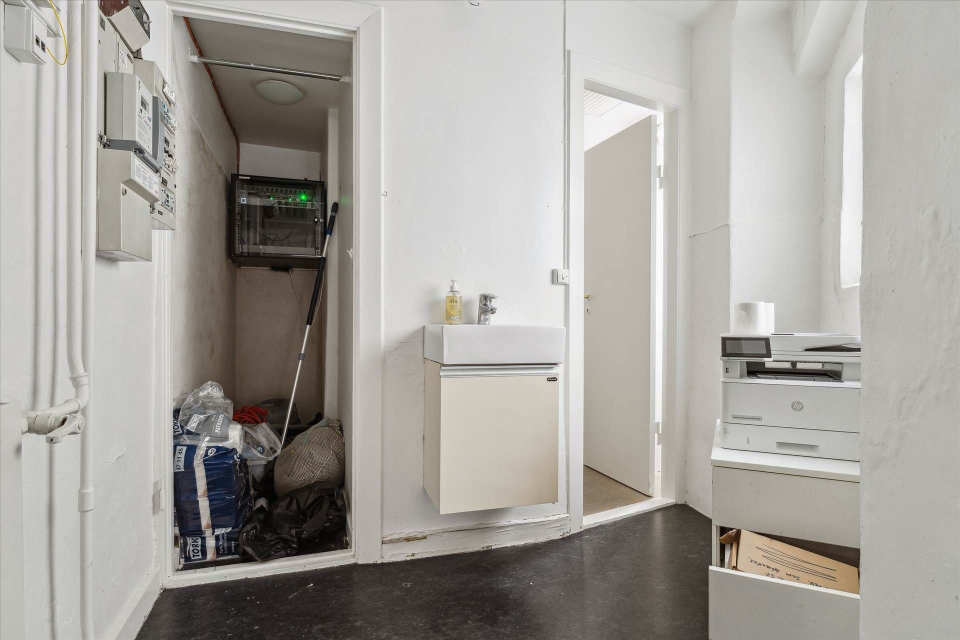 Hjørnerum med opvaskeblik, hvor der er en printer og papkasser i skabet, og der er et skab med udstyr på gulvet