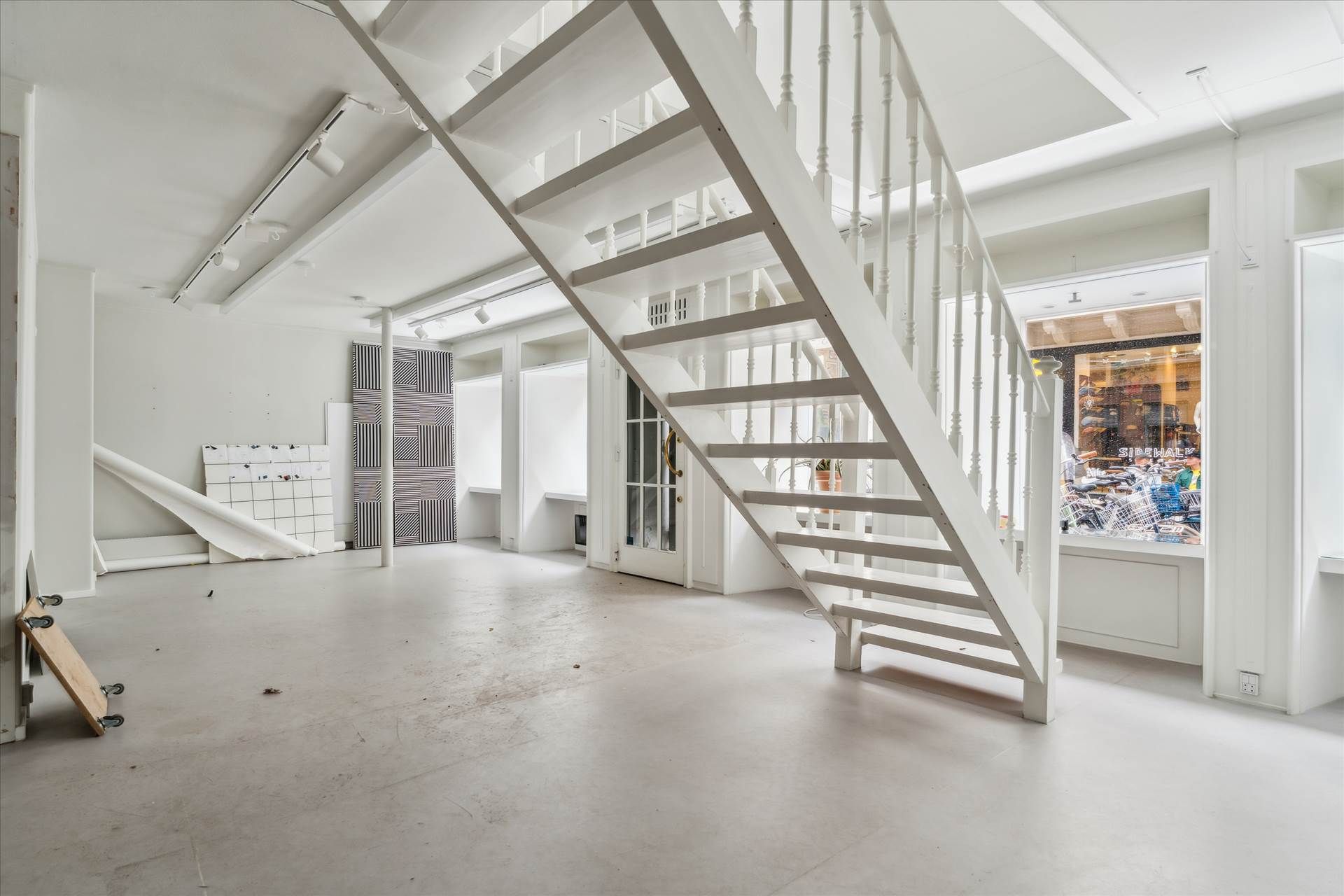 Et lyst og åbent rum med hvide vægge, loft og gulv samt en simpel trappe og et vindue med udsigt til butiksdisplayet.