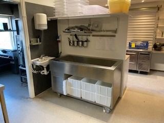 Catering køkken med stålvaskedsk og flere beholdere og sager i køkkenet, samt en sæbe dispenser og en håndsvampe ved