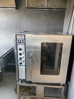 Køkken med en stor kommerciel ovn placeret på et metal stativ med flere knapper og en åben dør