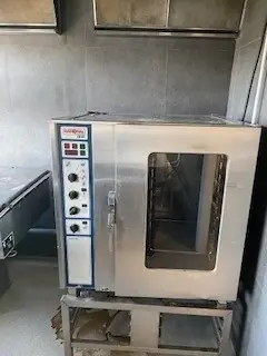 Køkken med en stor kommerciel ovn placeret på et metal stativ med flere knapper og en åben dør