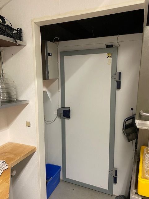 Et køkken med åben dør og en hvid kasse samt en blå kurv og en træhylde med glas og plast