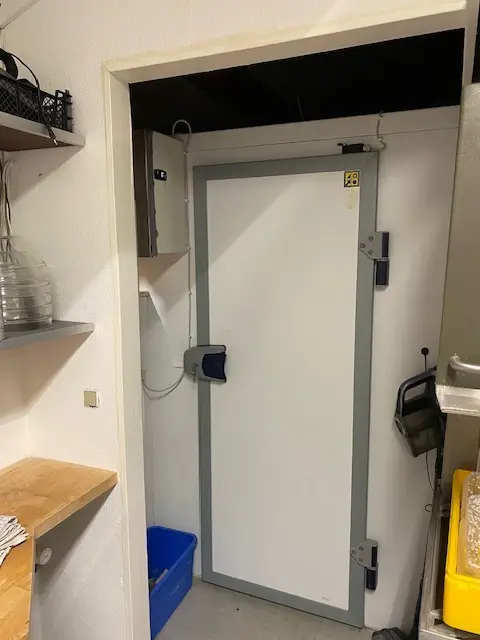 Et køkken med åben dør og en hvid kasse samt en blå kurv og en træhylde med glas og plast