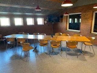 Et mødelokale med lysegult panel, lysekroner, tre vinduer, bord og stole, tv og en projektor på væggen