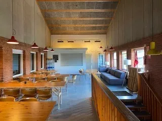 Lyserøde lamper hænger fra loftet, og der er flere borde og stole i rummet. Der er også en sofa og et skrivebord.