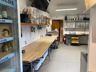 Et catering køkken med træbord, hylder med forskellige produkter, og lagerrum med diverse materialer, samt en lukket dør og