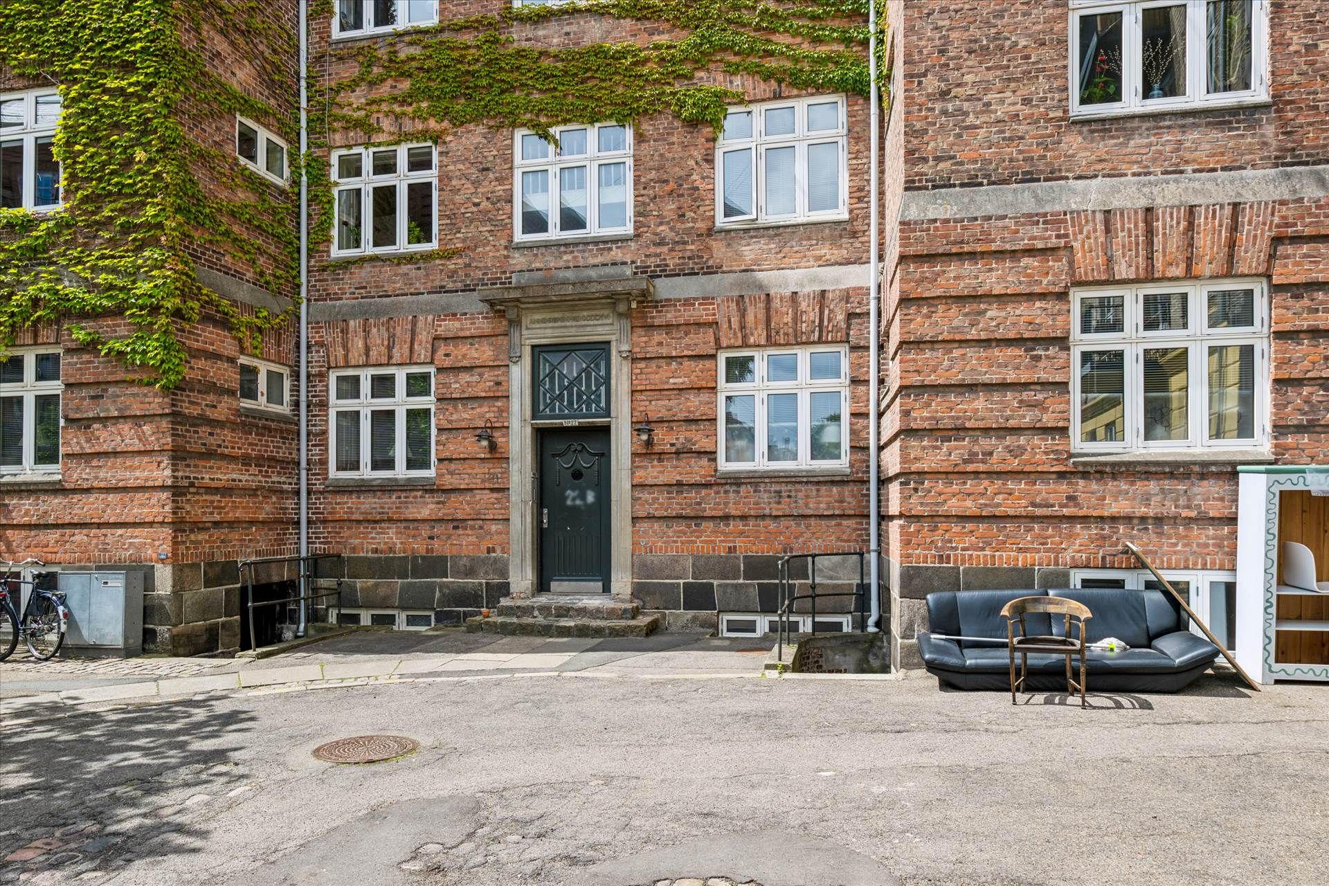 En gammel murstensbygning med grønne planter og flere vinduer, udenfor står en stol og en sofa samt en cykel