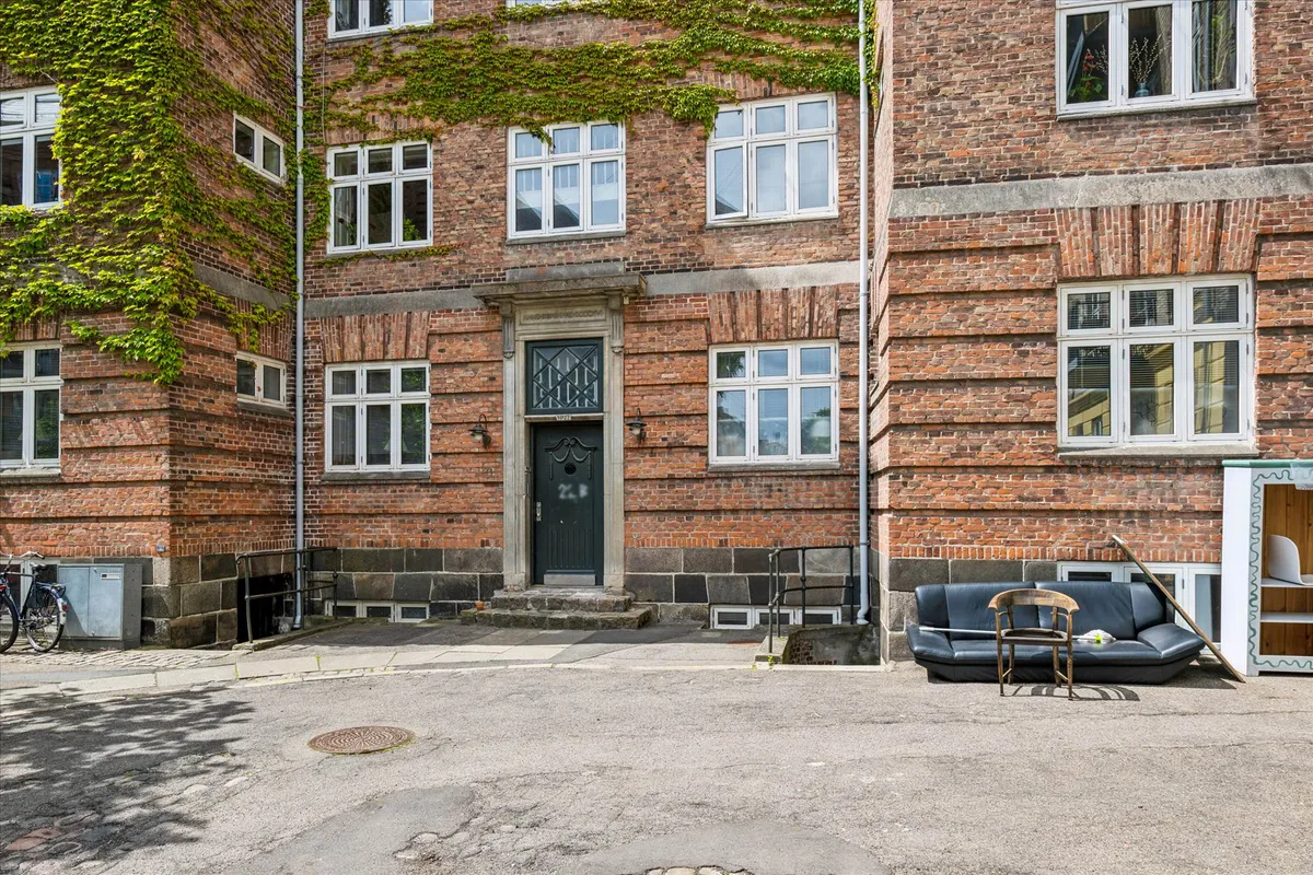 En gammel murstensbygning med grønne planter og flere vinduer, udenfor står en stol og en sofa samt en cykel