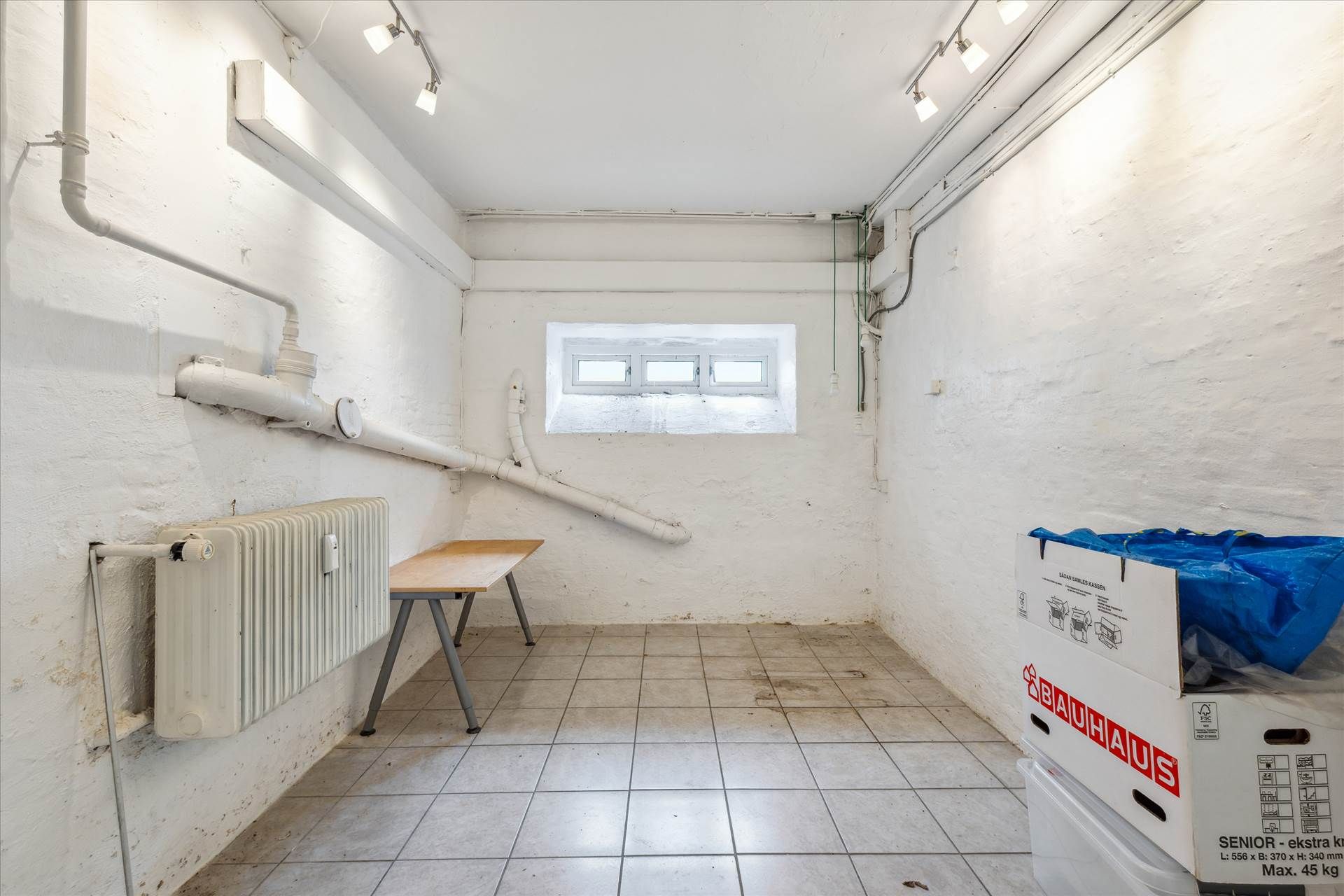 Et lagerrum med en radiator, et bord, et vindue og to bokse med Bauhaus-mærker og en blå pose