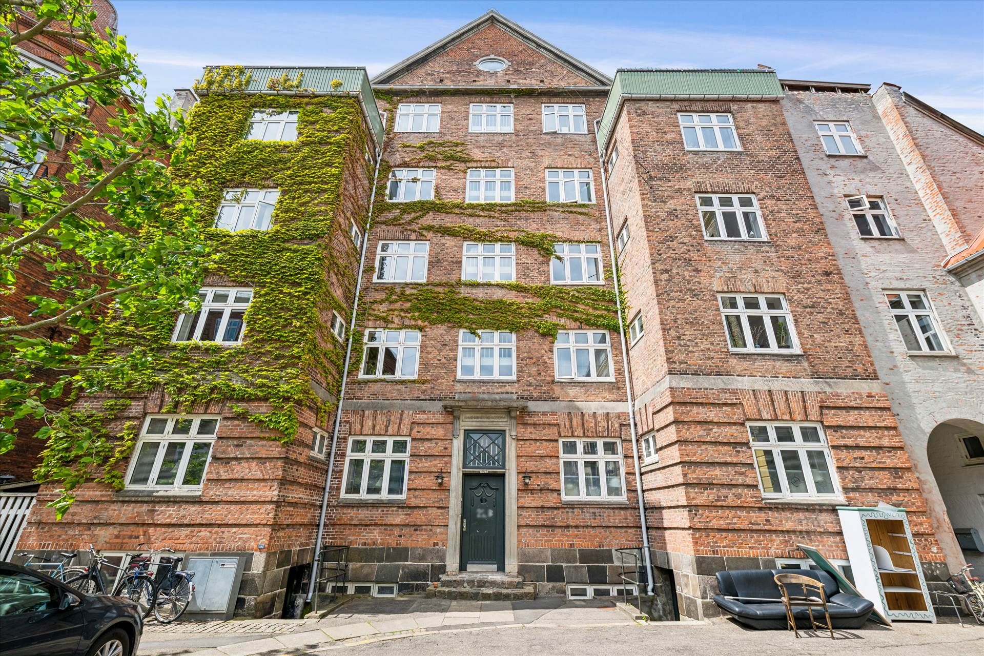I Frederiksberg står et gammelt rødt murstenshus med krybende planter og flere vinduer. Parkerede cykler og en bil uden for.