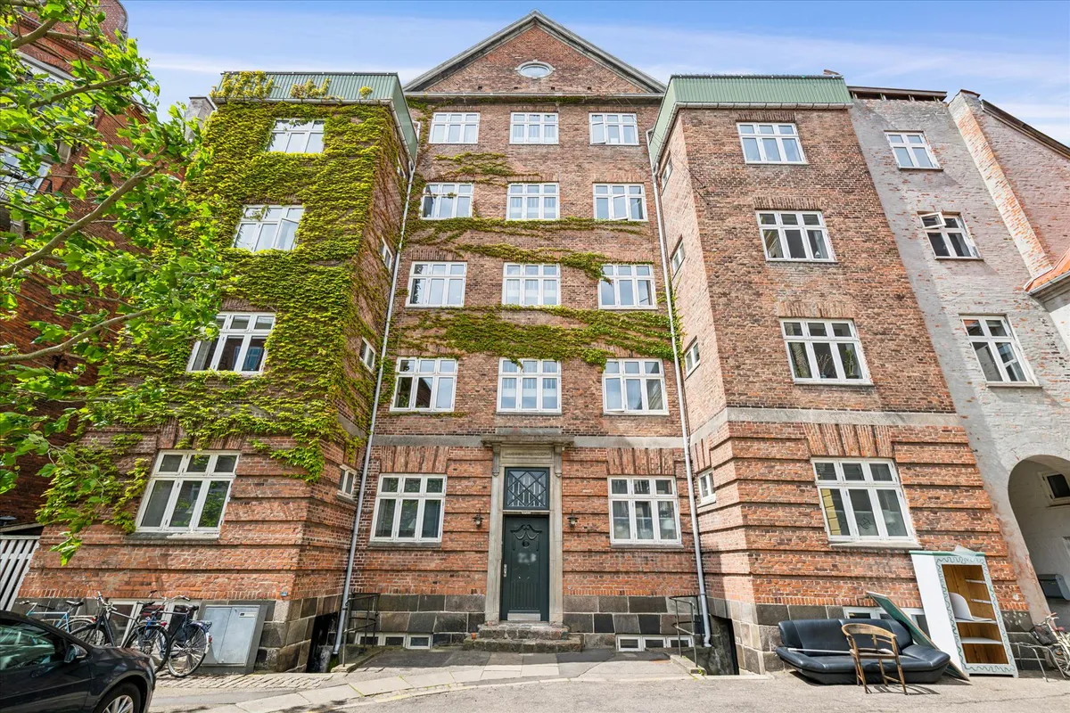 I Frederiksberg står et gammelt rødt murstenshus med krybende planter og flere vinduer. Parkerede cykler og en bil uden for.