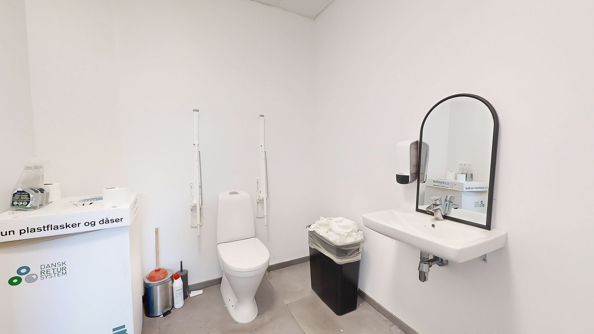 Rent og renset toilet med håndvask og spejl med håndsæbe og toiletpapir. Der er en skraldspande, rengøringsprodukter og