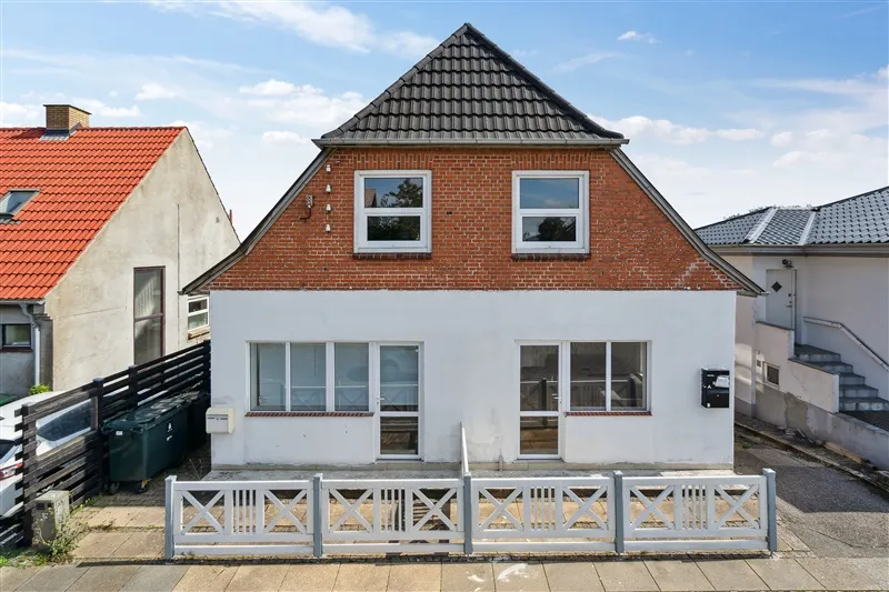 I baggrunden er et rødt tag og en gul sidevæg på et hus. Foran er et hvidt hus med røde mursten og store ruder. Der er et