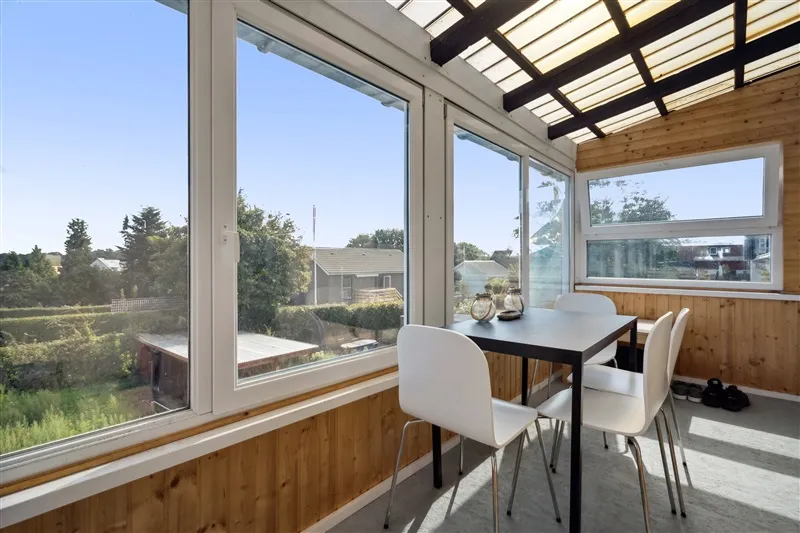 Vindueskarmen med træ og glas. Bord og stole med udsigt til have med træer og hus i baggrunden.