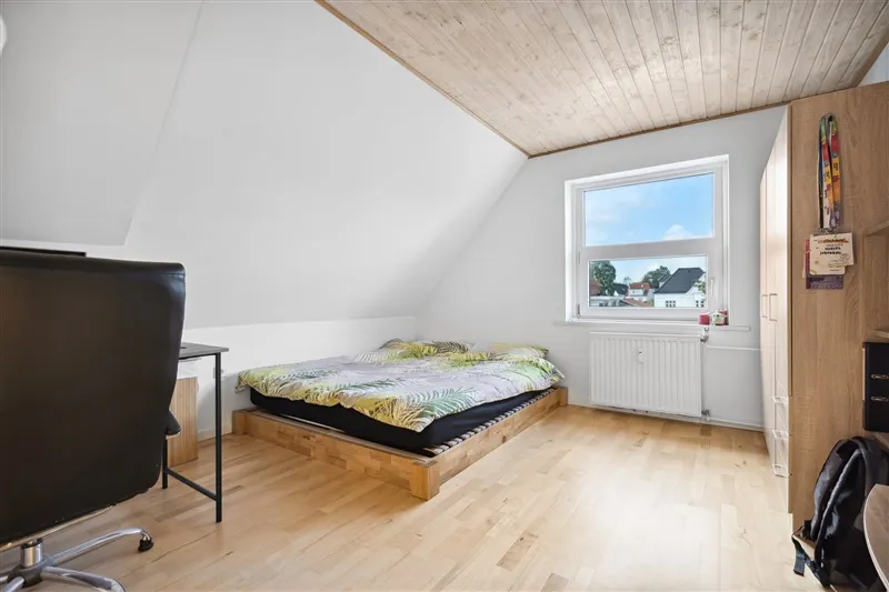 Et rum med højt loft og hårdt trægulv, et dobbeltseng med lyse dynebetræk og et skrivebord med en stol og en rygsæk