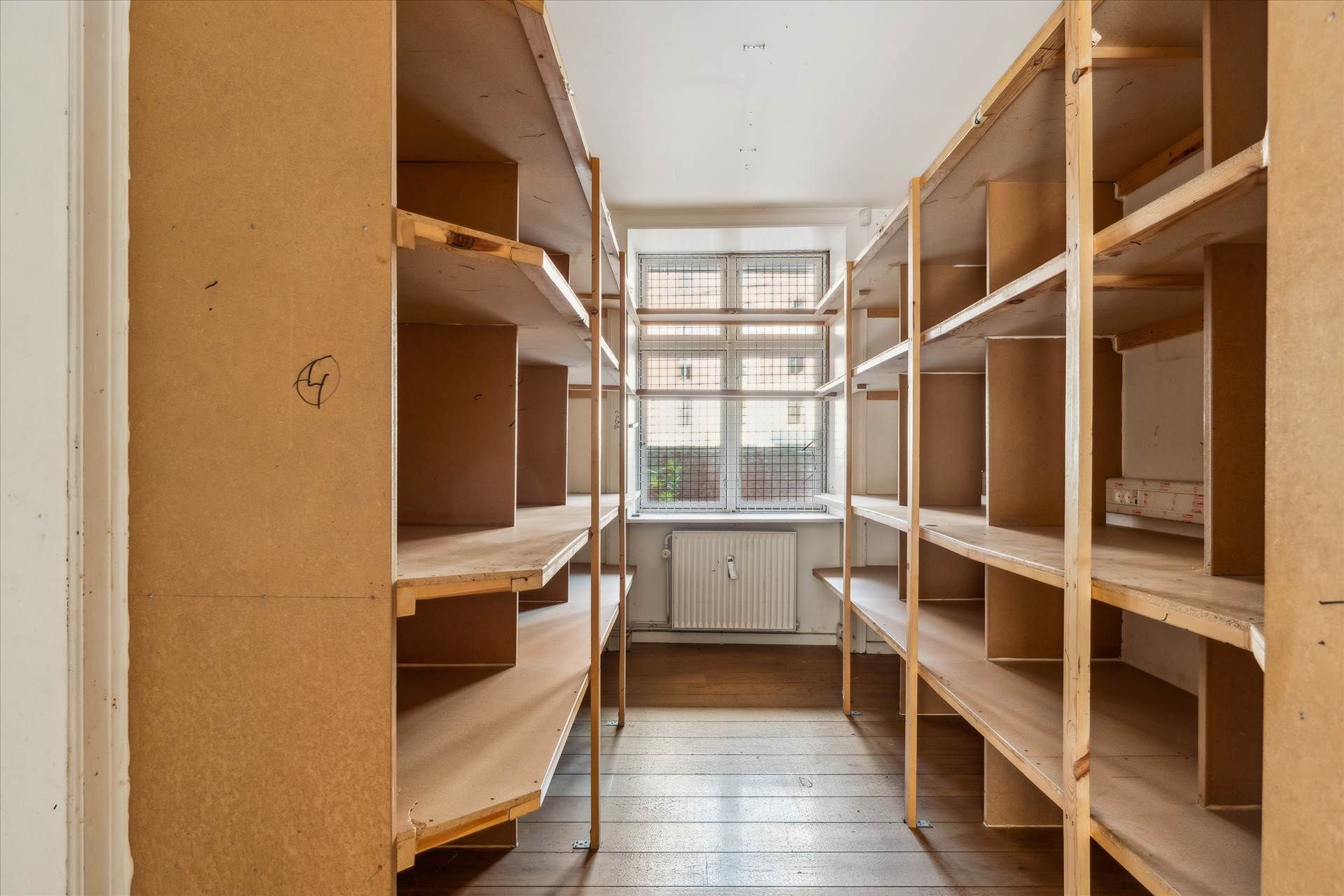 Lagerrum med to rækker af træhylder, en radiator og vinduer med sikringsgitter i baggrunden