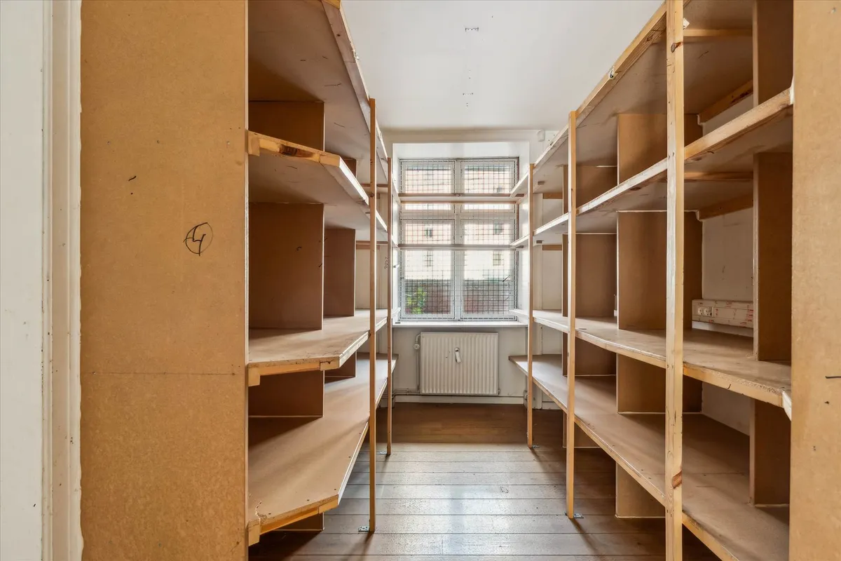 Lagerrum med to rækker af træhylder, en radiator og vinduer med sikringsgitter i baggrunden