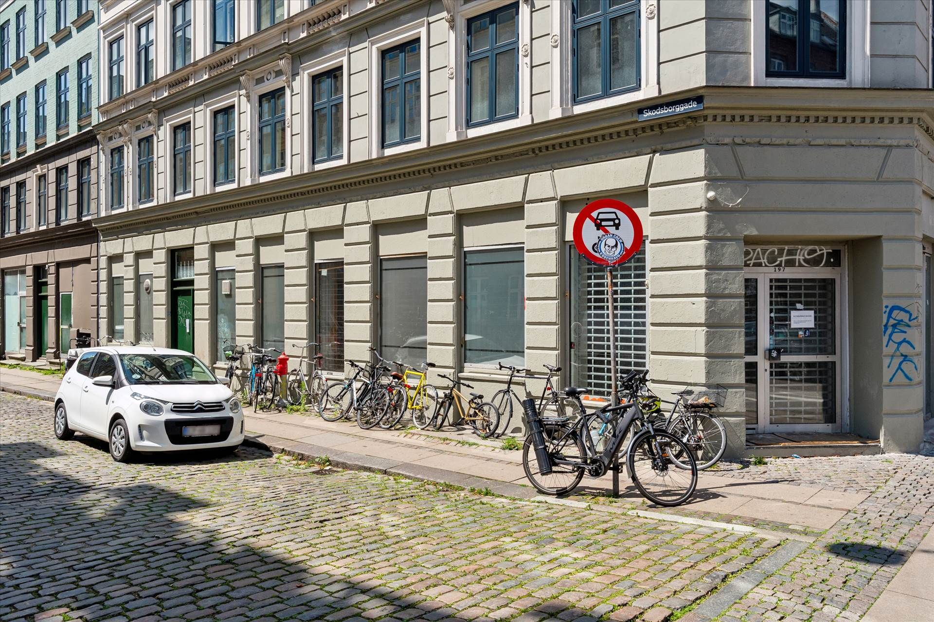 Hjørnebygning med adgangsforbud for biler og cykler parkeret på fortauet, samt en hvid bil parkeret foran.