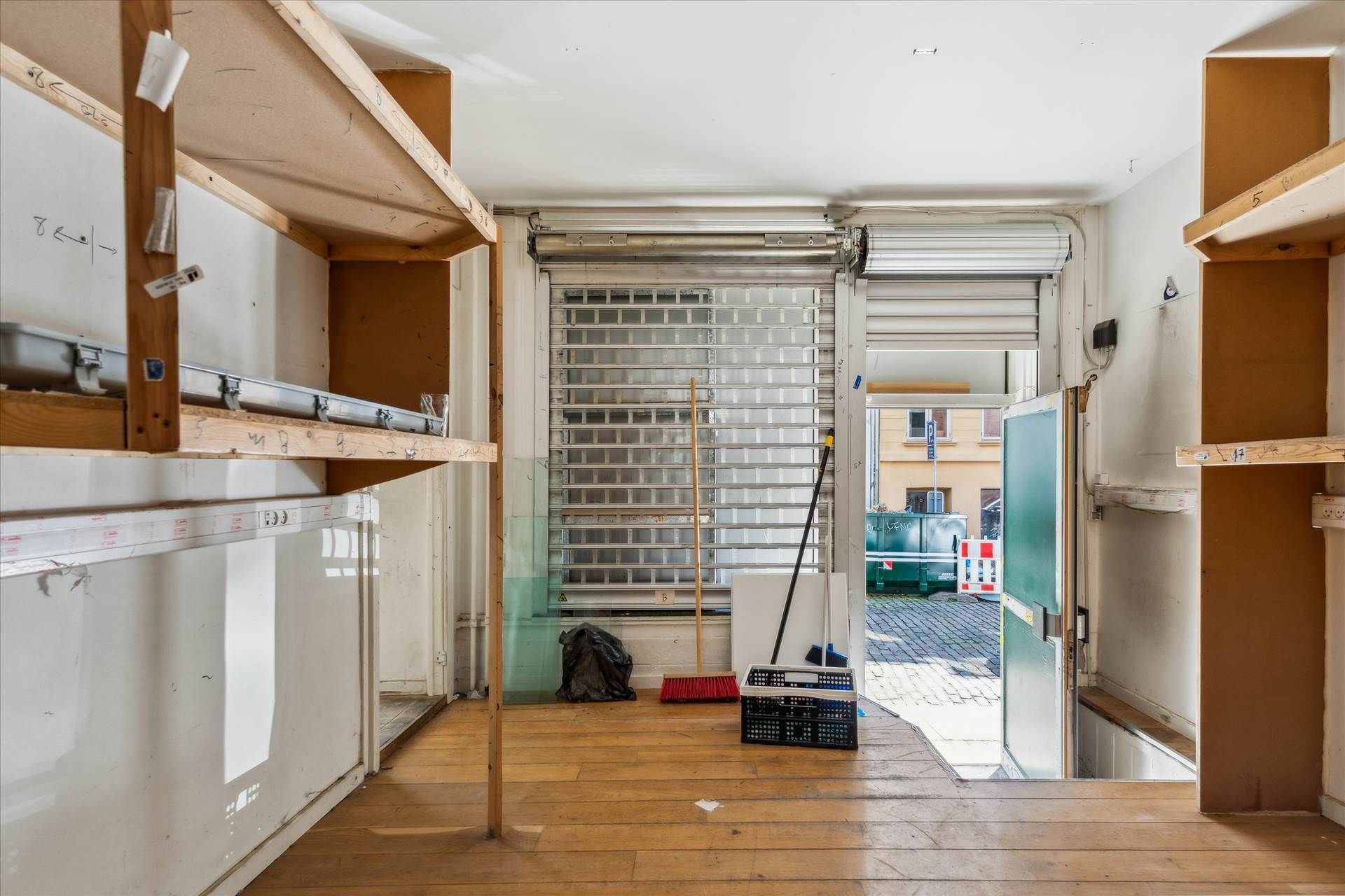 Tomt og lukket erhvervslejemål med store vinduer og trægulv. Træhylder og opbevaringskurve står klar til brug