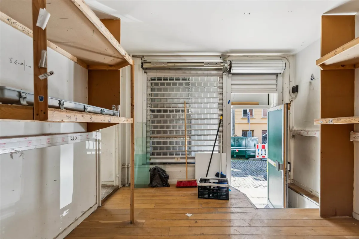 Tomt og lukket erhvervslejemål med store vinduer og trægulv. Træhylder og opbevaringskurve står klar til brug