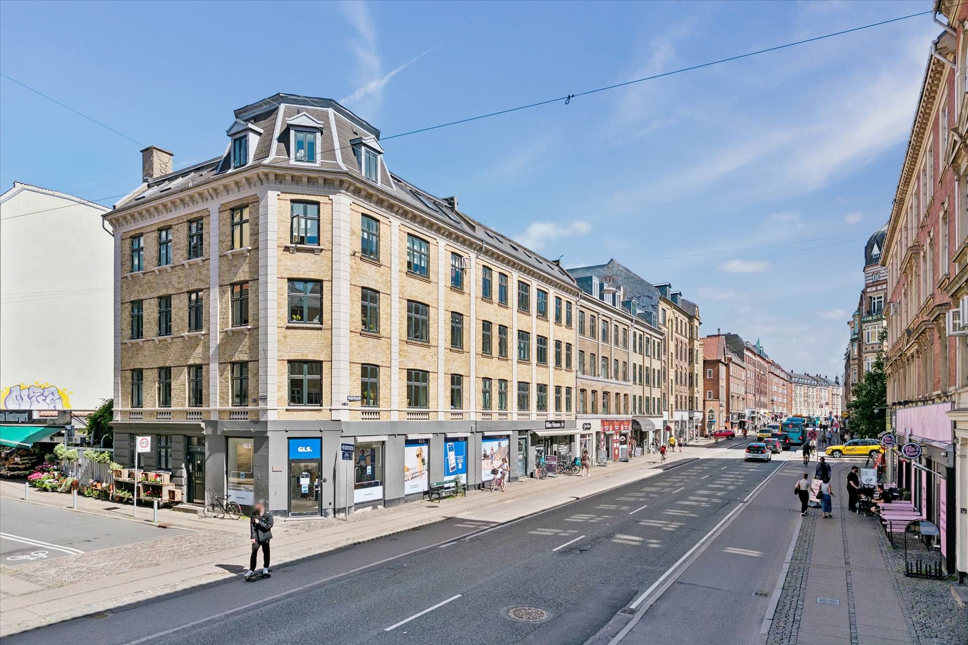 Butik med hjørnebygning, hvor folk går forbi og en cyklist kører på Amagerbrogade med en bus og biler i baggrunden