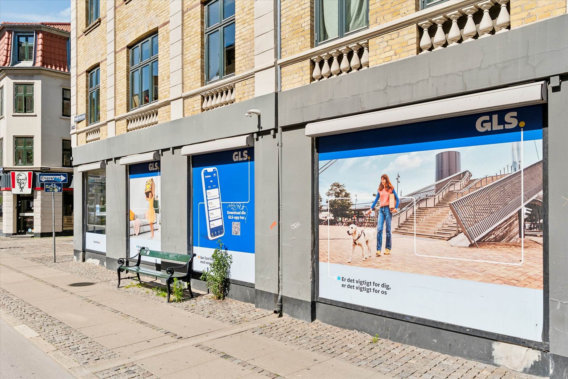 En butik med reklameskilte for GLS på siden af en gågade, med en kvinde og hund i reklamen, samt en kvinde på en sofa i en