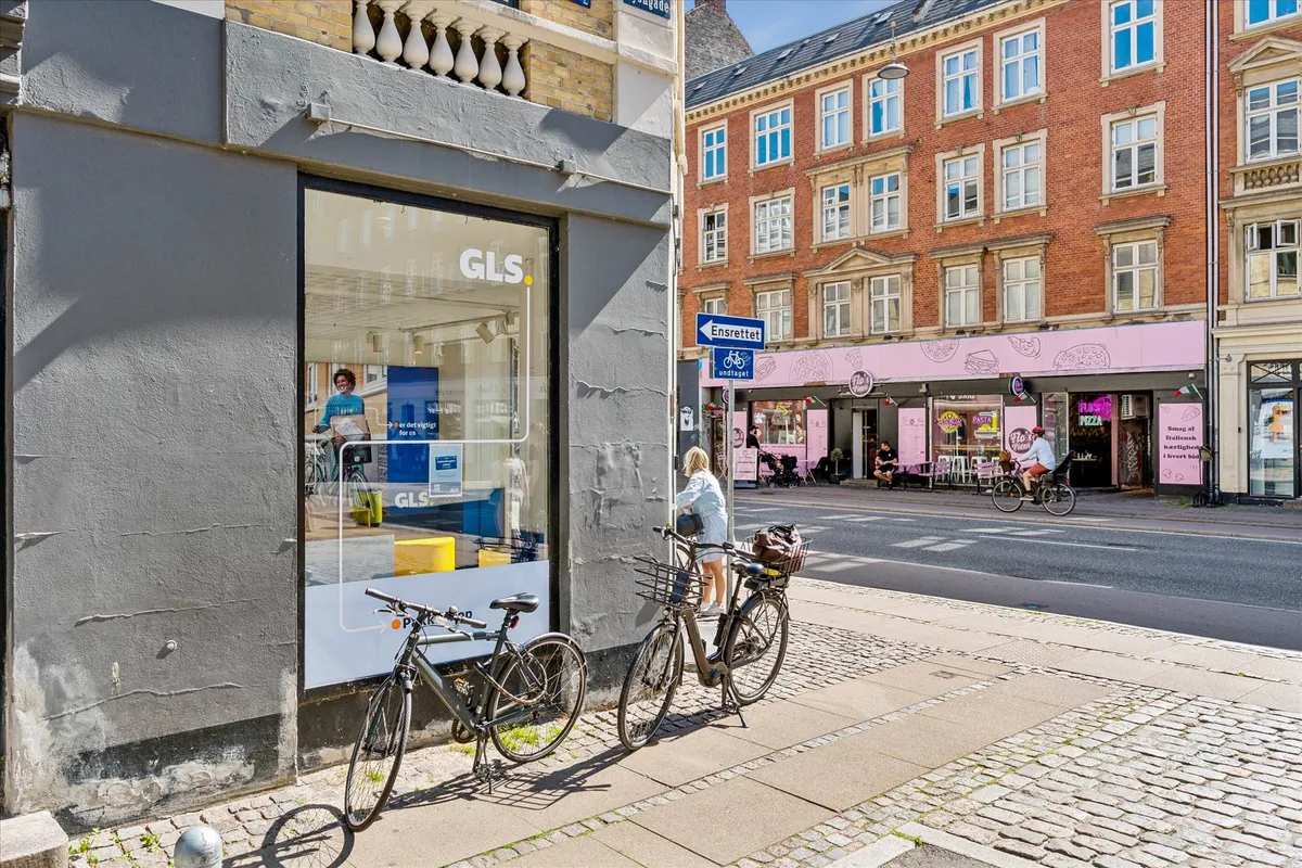 To cykler er parkeret udenfor en butik med navnet GLS, hvor en voksen og et barn står. En cykel er parkeret på hjørnet, og