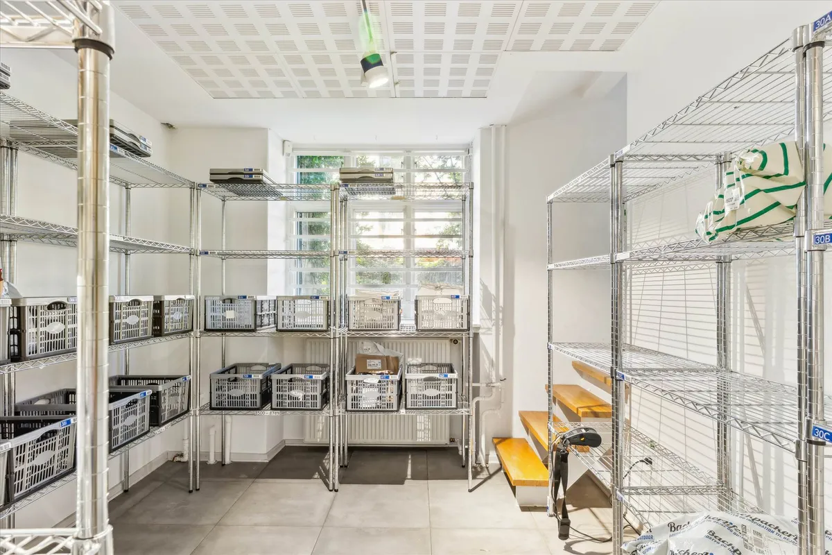 Foto af et køkken med høje, metal hylder og bøsser med etiketter, hængende loftslampe, vinduer med gardiner og en træbænk