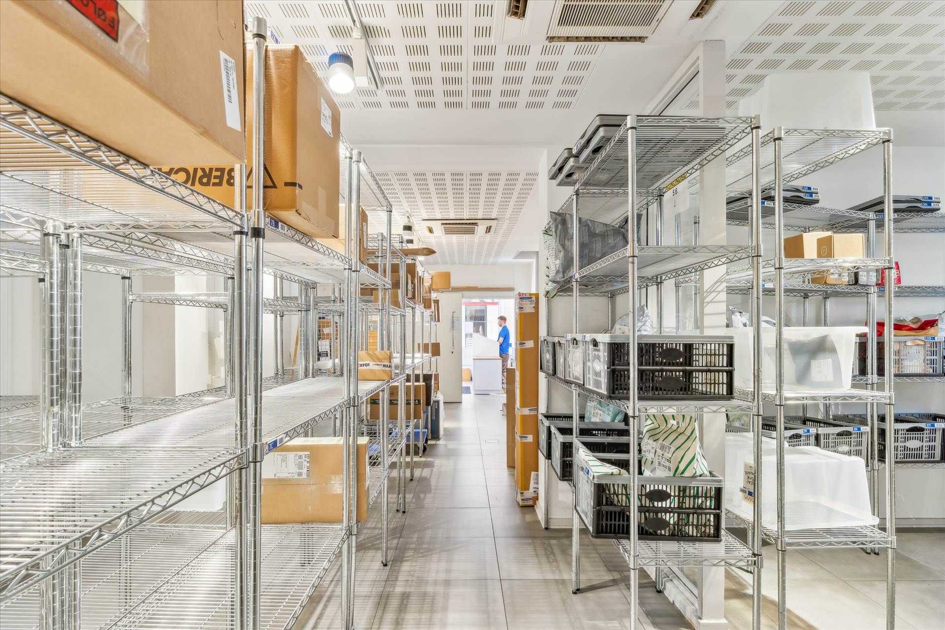 Lager med flere lagerhylder med både kasser og plastposer. Høje hylder og et åbent rum med et kryds på gulvet.
