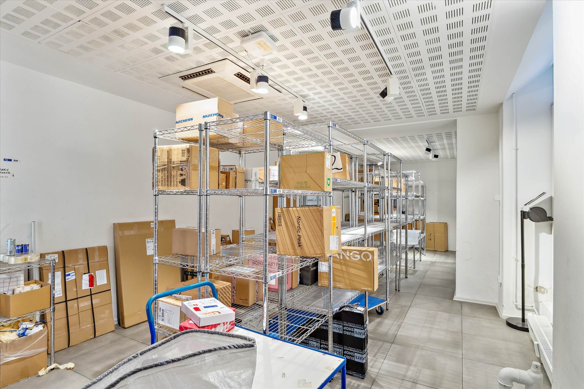 Lager med mange forskelige kasser og bokse samt højrekantede hylder. Der er flere lysekroner på loftet og gulvbelægning af