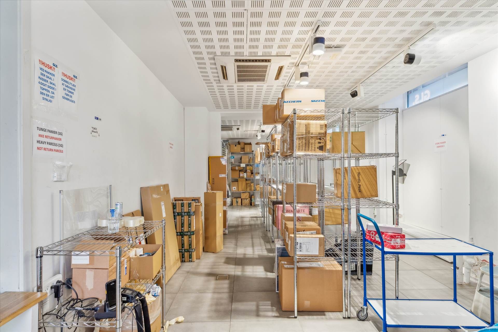 Lager med kartoner og emballagematerialer, og en rullebrod, stående i et stort rum med højt gulv og hvide vægge