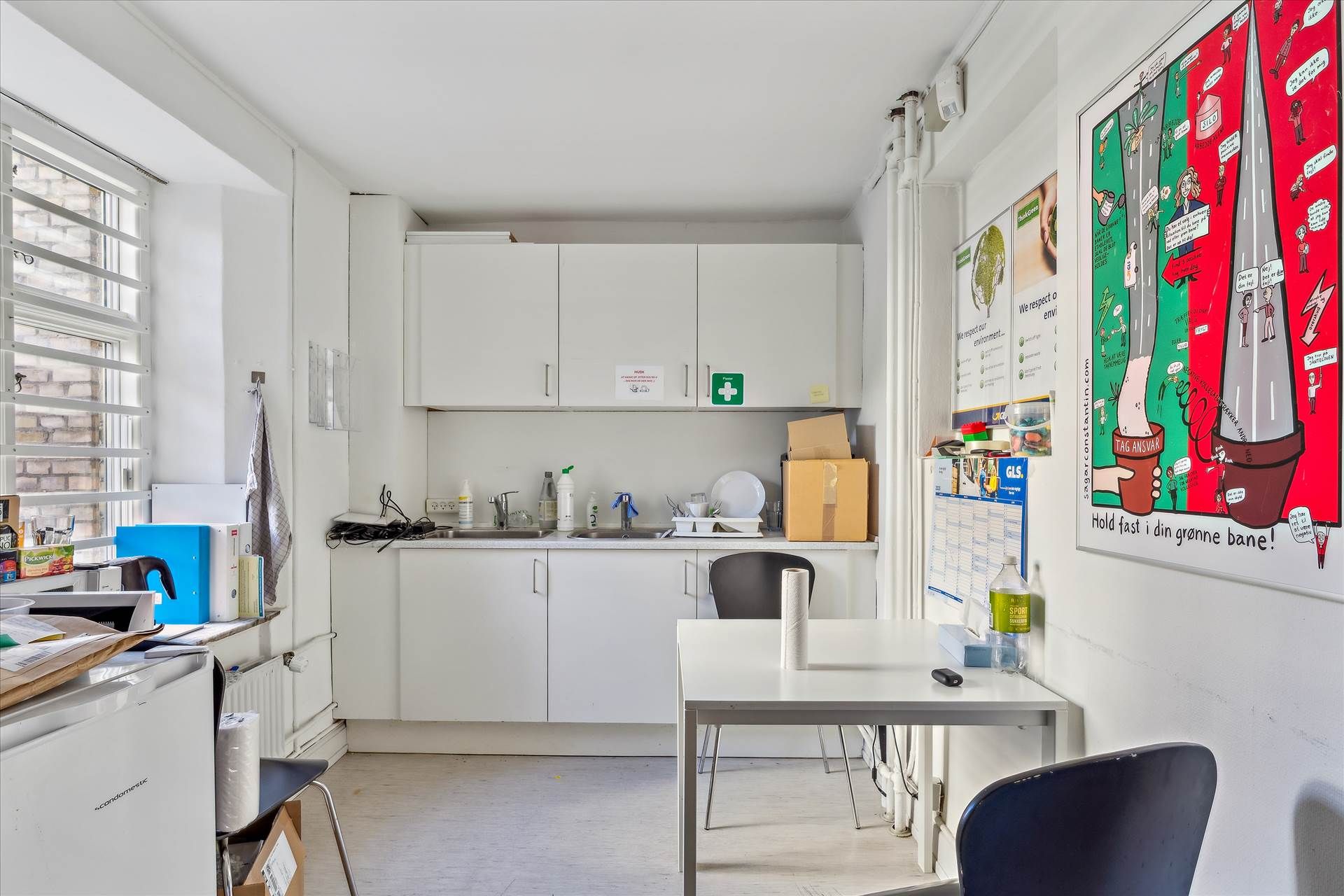 En stue med hvide vægge og højloft, med et køkken og en hvid bord med stoff på. Et køleskab og en mikrobølgeovn er placeret