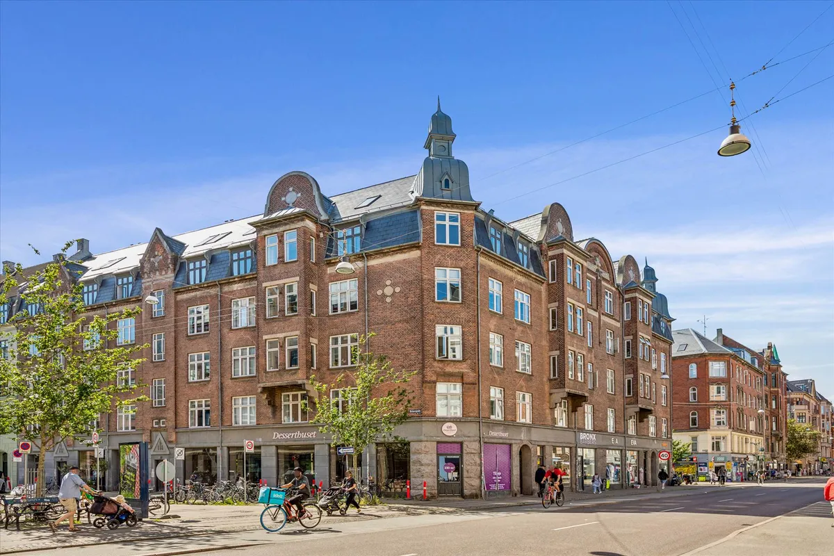 Forretningsbygning med mange vinduespartier og en fortovet gågade med cyklister og gående fodgængere på Nørrebrogade i