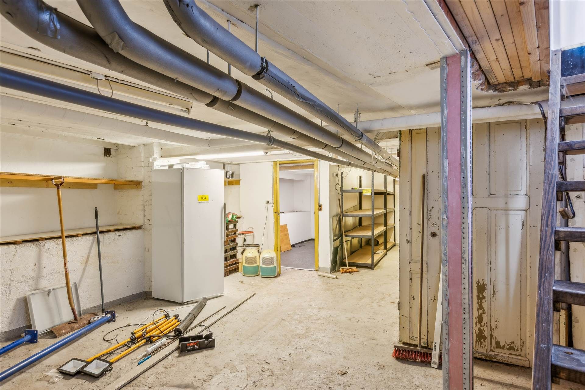 Lager med diverse værktøj og apparater, hvor en stor hvid apparatkasse står mod væggen og andre genstande som skovle og