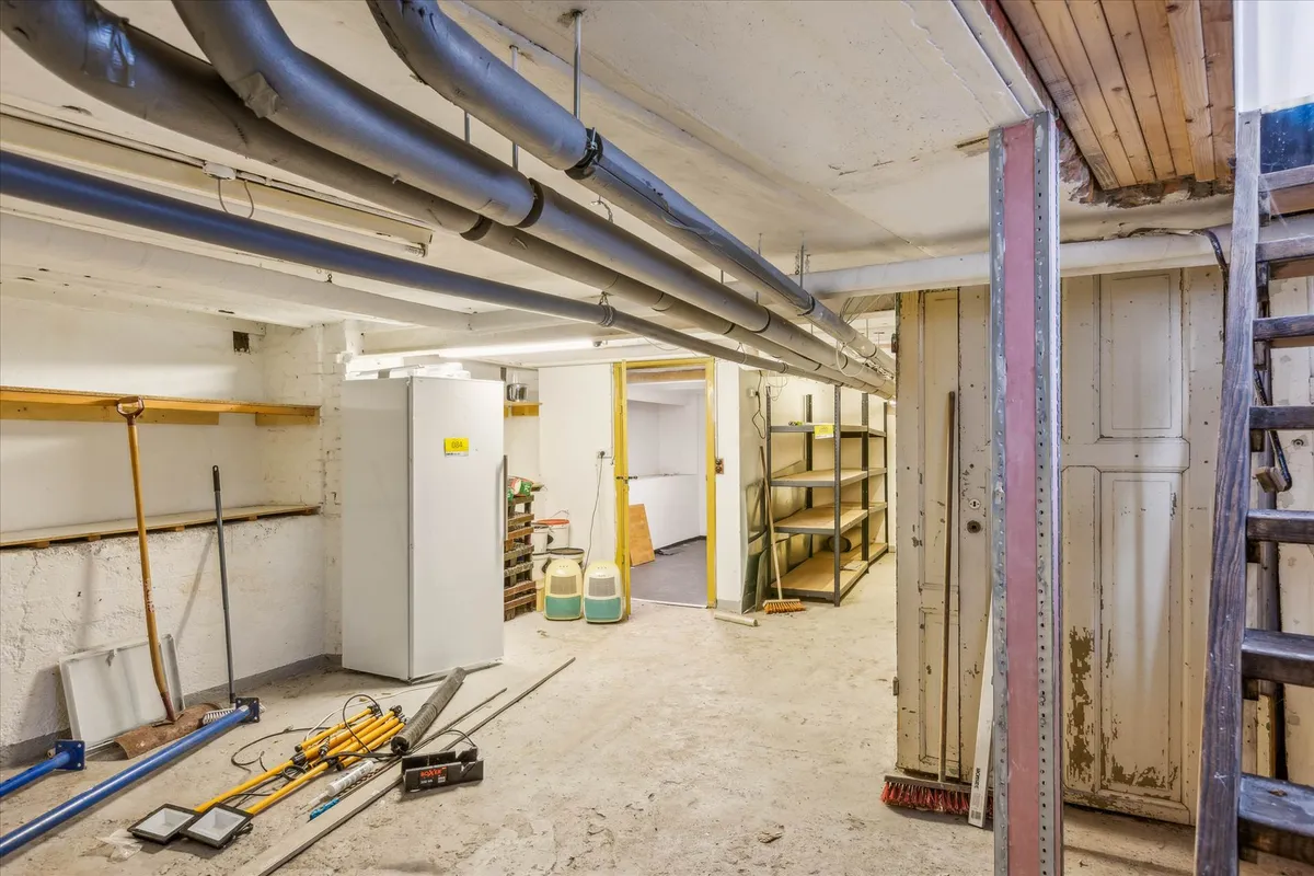 Lager med diverse værktøj og apparater, hvor en stor hvid apparatkasse står mod væggen og andre genstande som skovle og