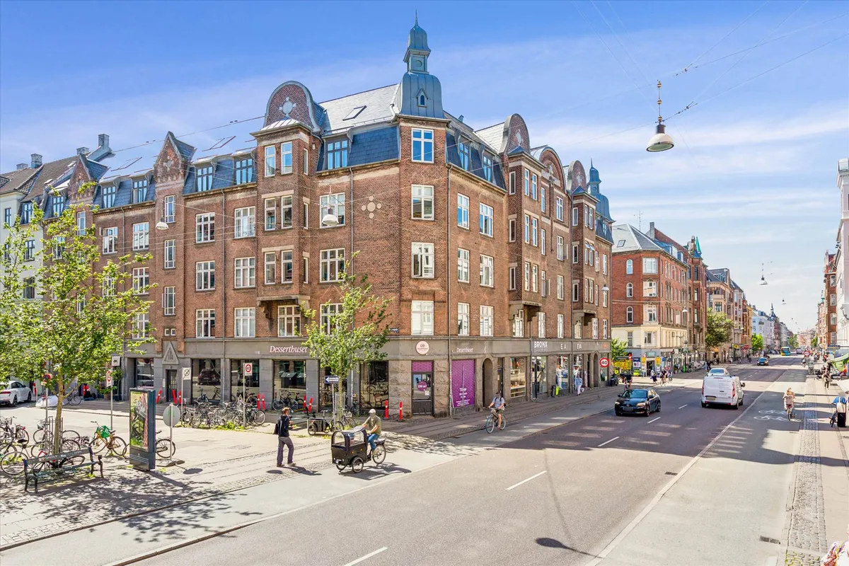 Mennesker går langs Nørrebrogade med biler og cykler. Store butikker med mange vinduer og forhøjede gader med trær og søjler