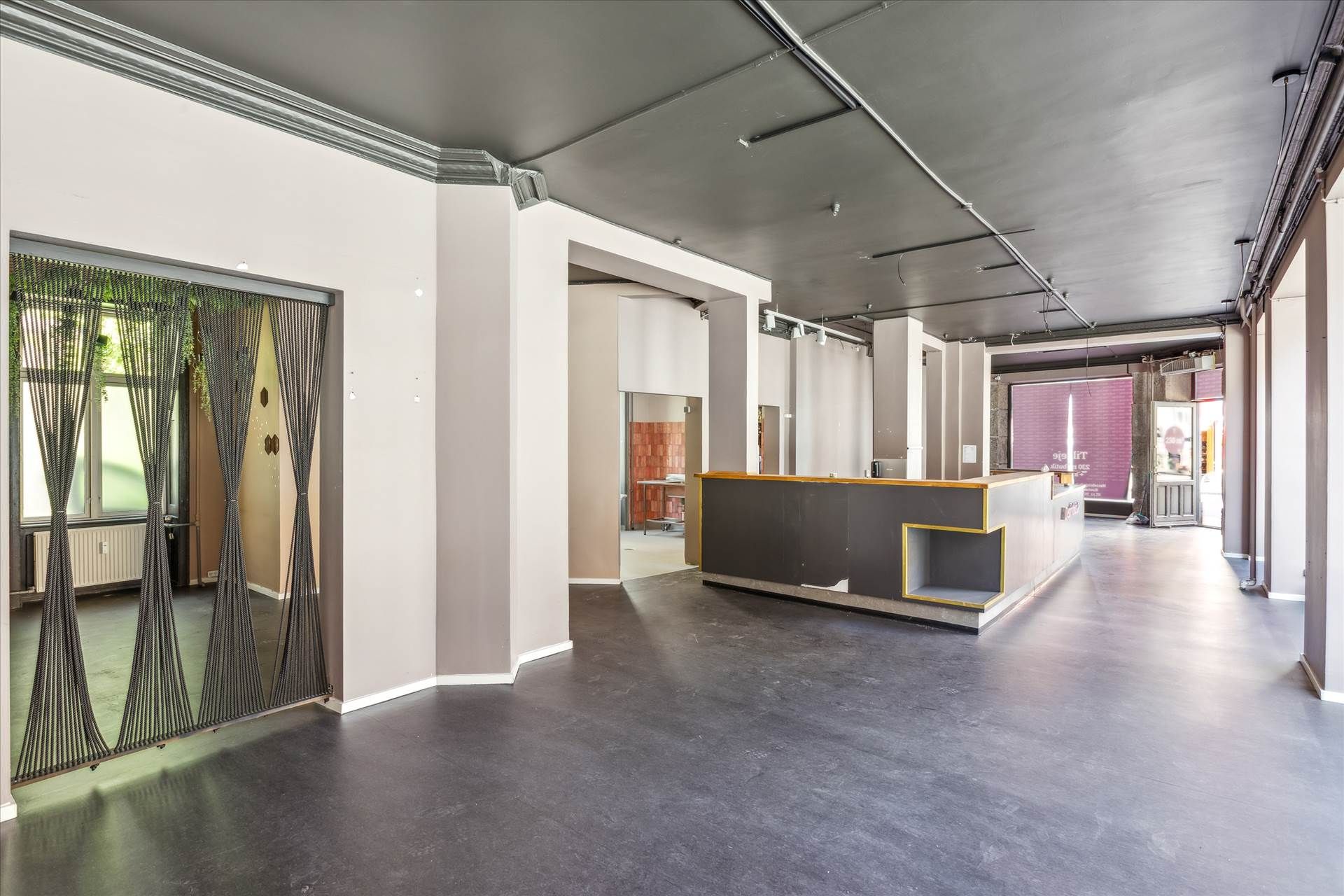 Et tomt lokale med højt til loftet, lysegulv, hvide vægge, en stærkt belyst bar, og et vindue med grønne plantager udenfor