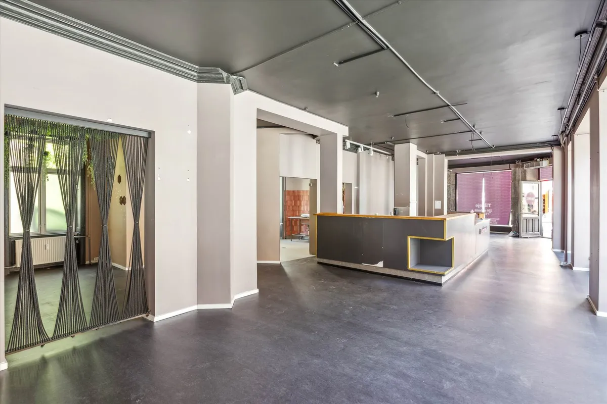 Et tomt lokale med højt til loftet, lysegulv, hvide vægge, en stærkt belyst bar, og et vindue med grønne plantager udenfor