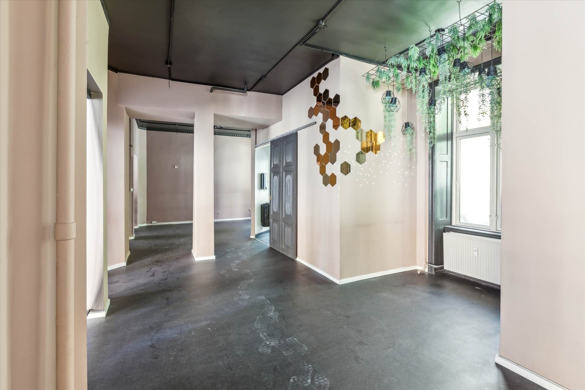 En tom gangvej med højt loft, gulvet er gul og lyse hjørner. Der er to døre og et gulv med en plante på væggen. Der er et